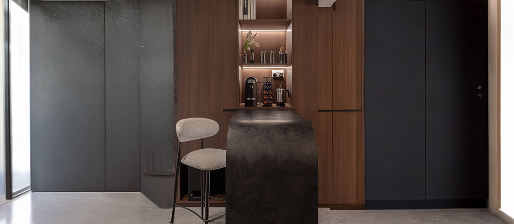Modern kitchen with curved dark island, gray bar stool, wood cabinetry, and accent-lit shelves with decor.
